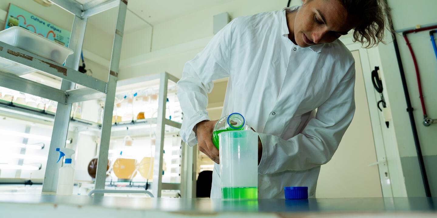 Personne en blouse blanche dans un laboratoire en train de faire un transfert de liquide verte entre deux contenants.