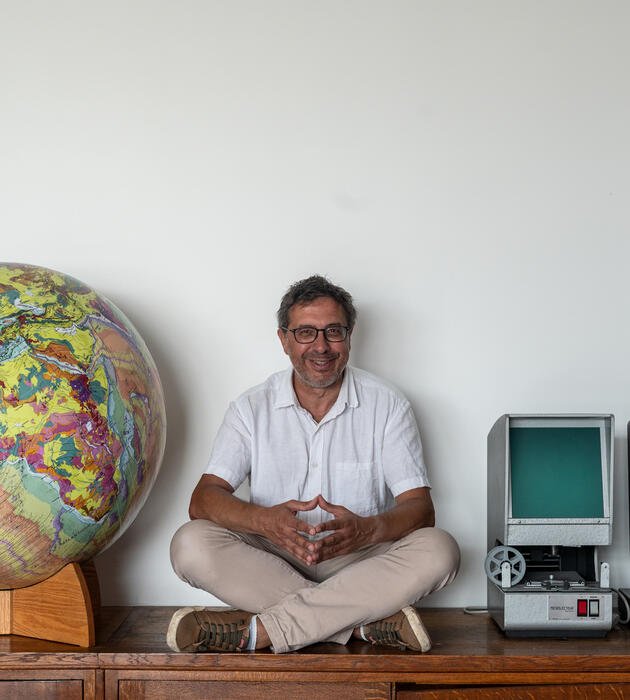 Homme assis sur un meuble avec à sa gauche deux ordinateurs et à sa droite un globe.
