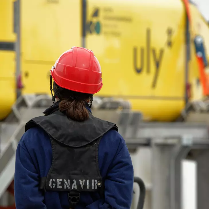 Personne de dos avec un casque rouge et un gilet de sauvetage estampillé "Genavir" devant un engin sous-marin sur le pont d'un bateau.